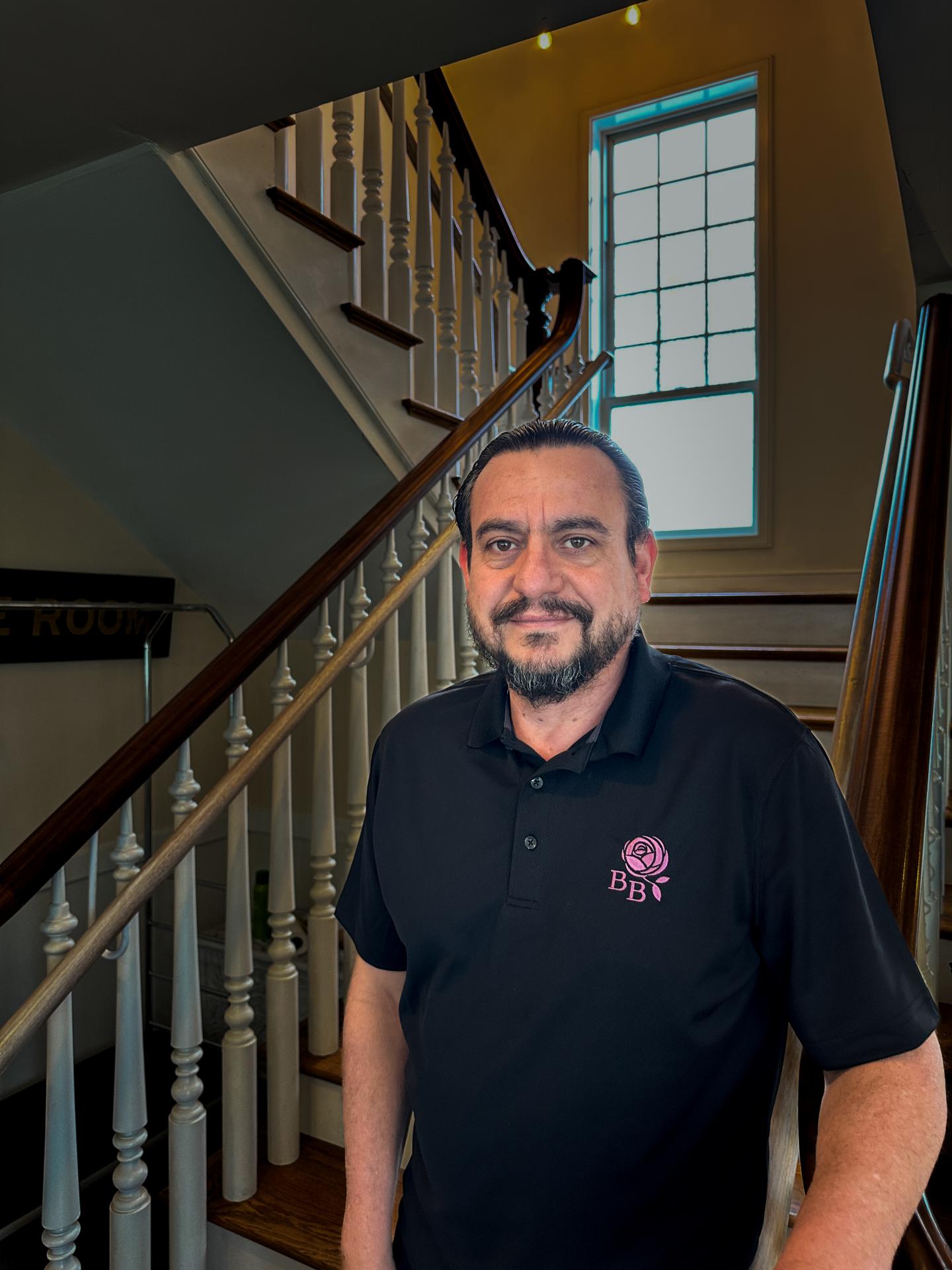 Man standing by a staircase, window in the background, wearing a black collared shirt.
