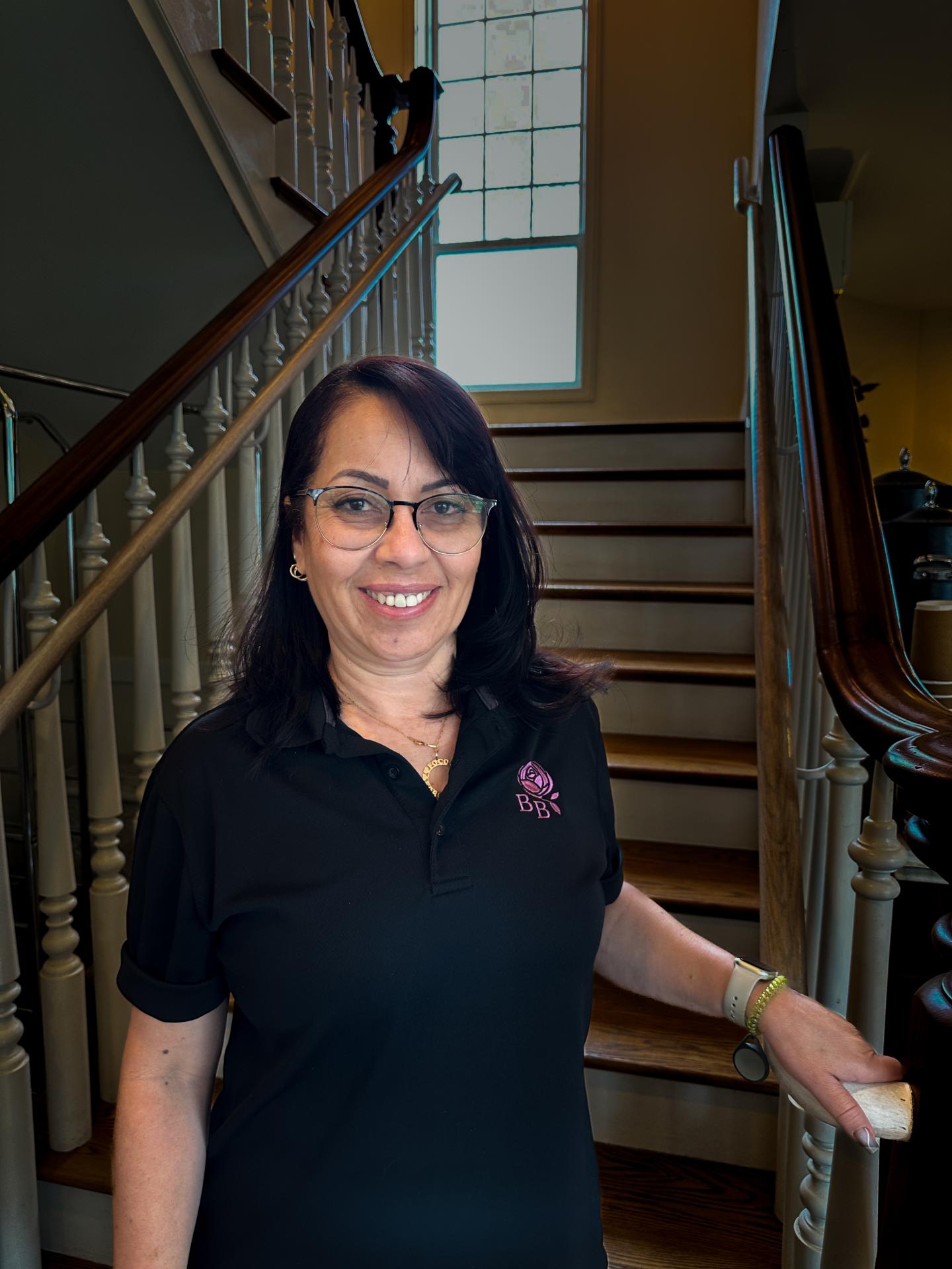 Woman smiling in front of a staircase, wearing glasses and a black polo shirt.