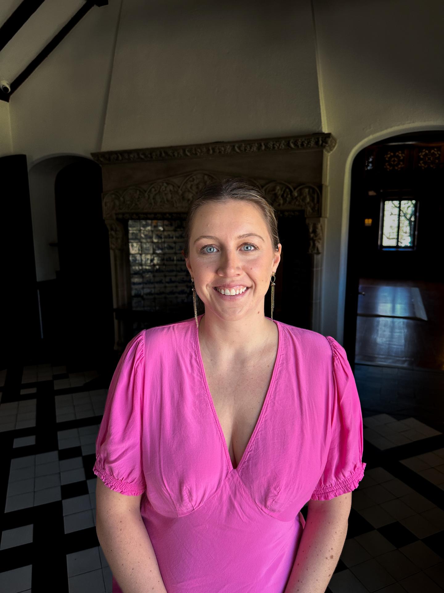 Smiling person in a pink shirt standing indoors with dark flooring and archway.