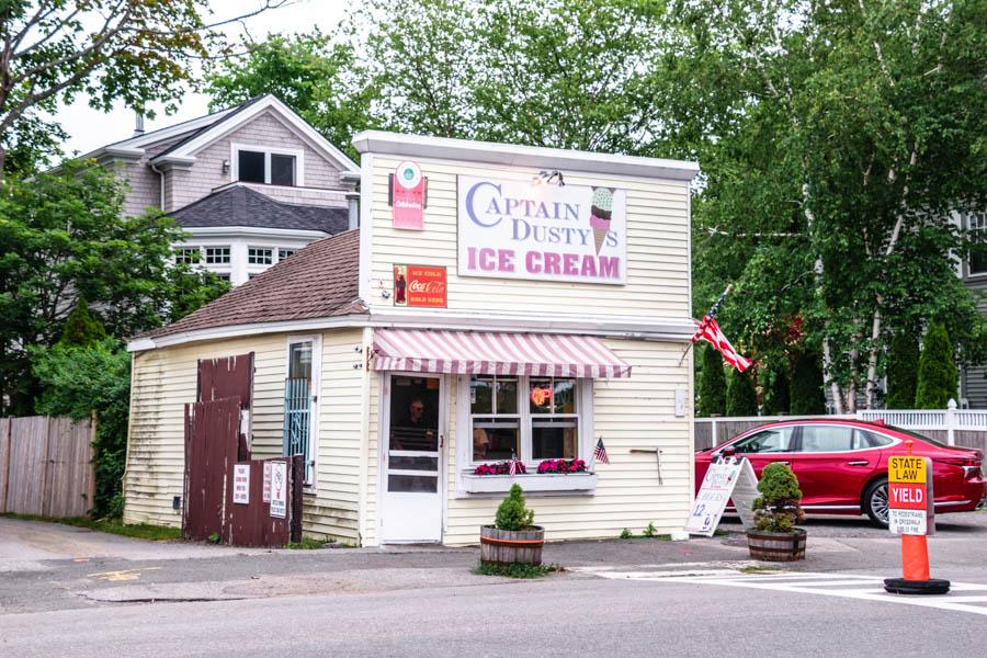 Small ice cream shop with yellow siding, awning, flag, and parked red car.
