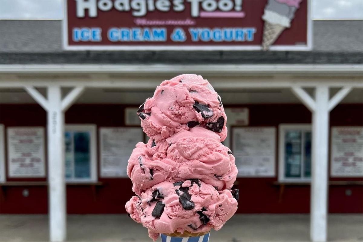 Triple scoop of pink ice cream with chocolate chunks, shop in background.