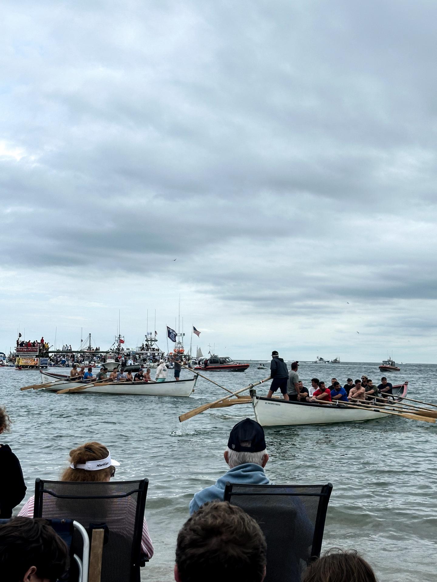 Rowboats with people in the sea under a cloudy sky, viewed from the shore.