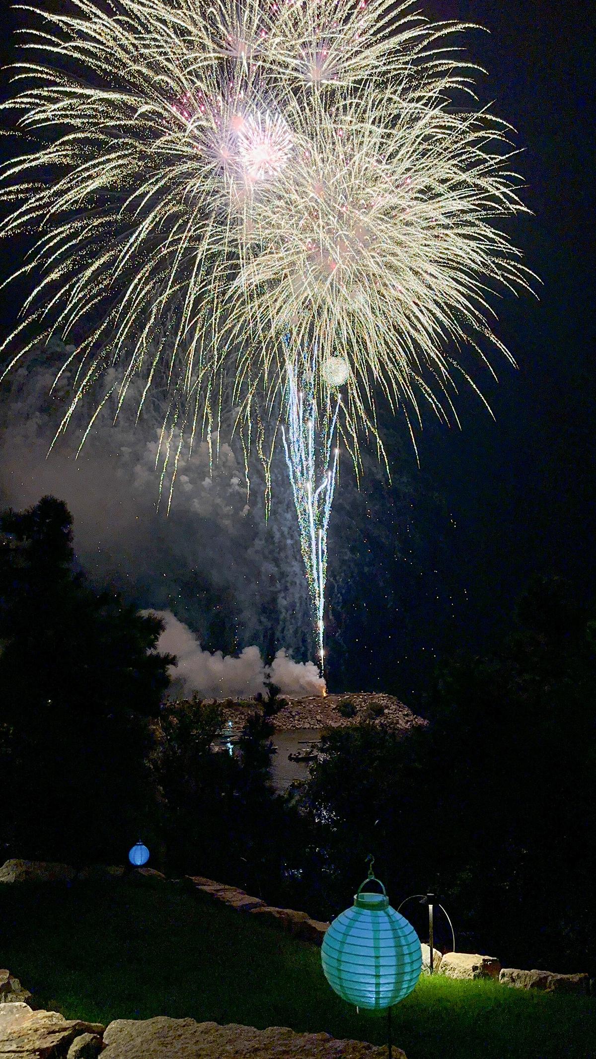 Fireworks burst in the night sky above silhouetted trees and glowing lanterns.