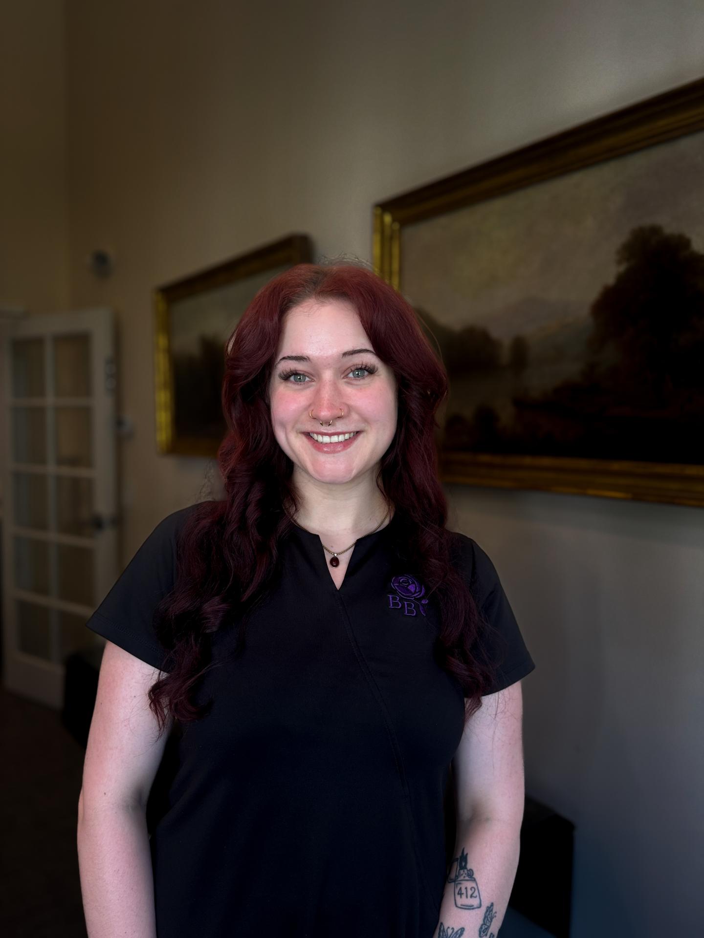 Smiling woman with long hair in a dark polo shirt, standing indoors by framed paintings.