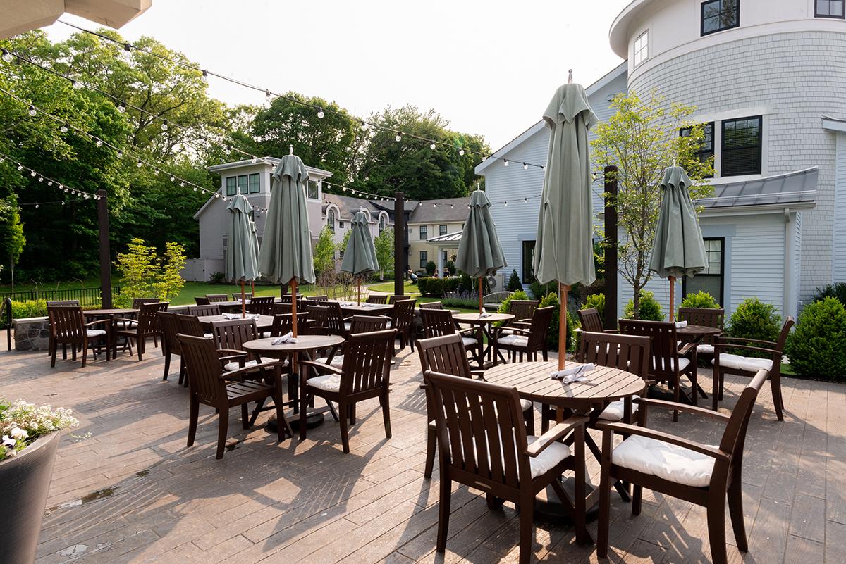 Outdoor café patio with wooden tables, chairs, and umbrellas, surrounded by greenery.