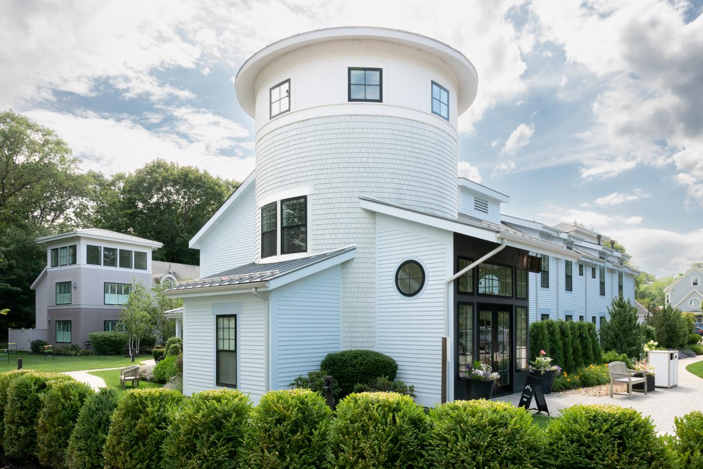 Modern house with circular tower, white siding, and lush greenery under a cloudy sky.