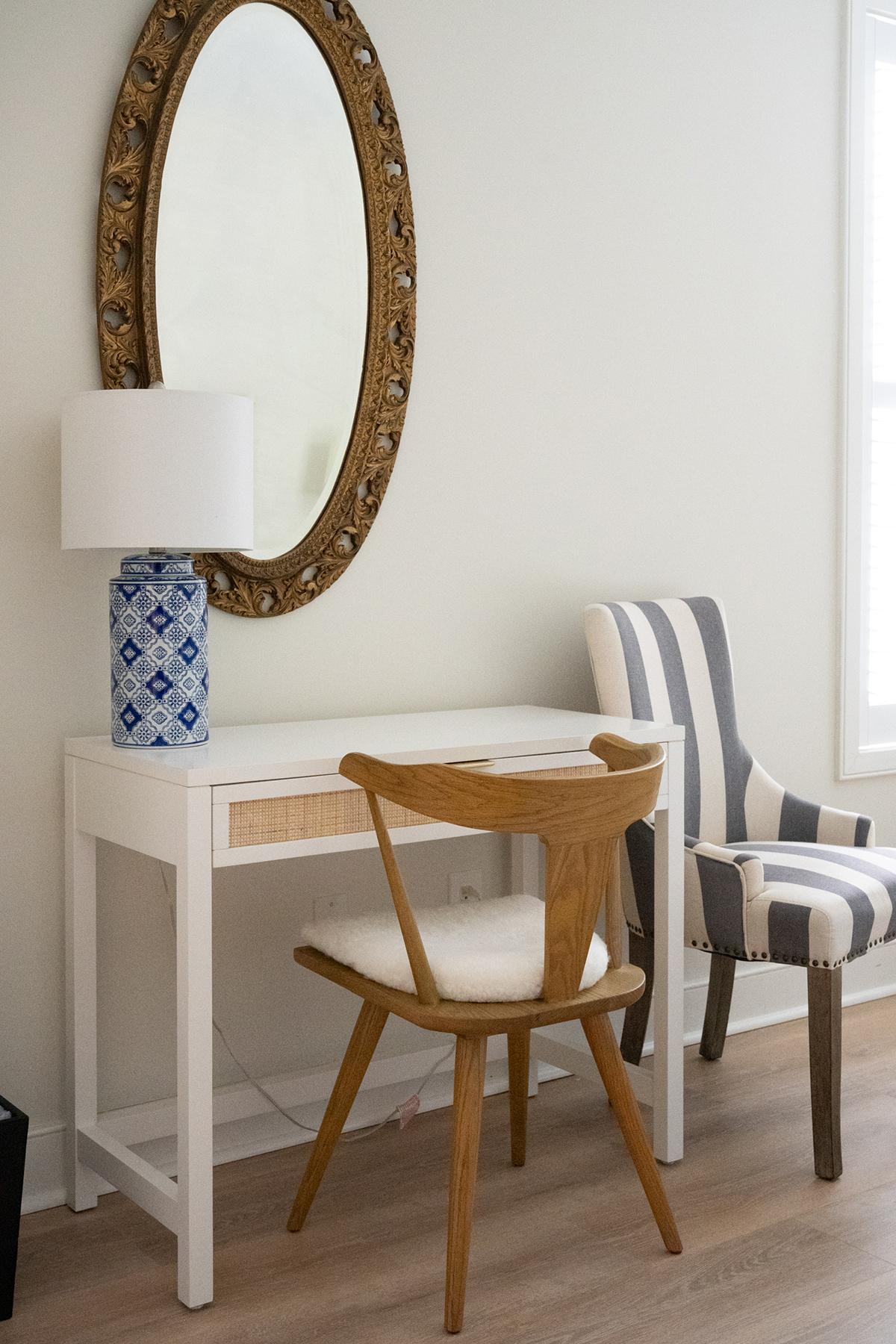 Desk with oval mirror, striped chair, and blue lamp in a bright room.