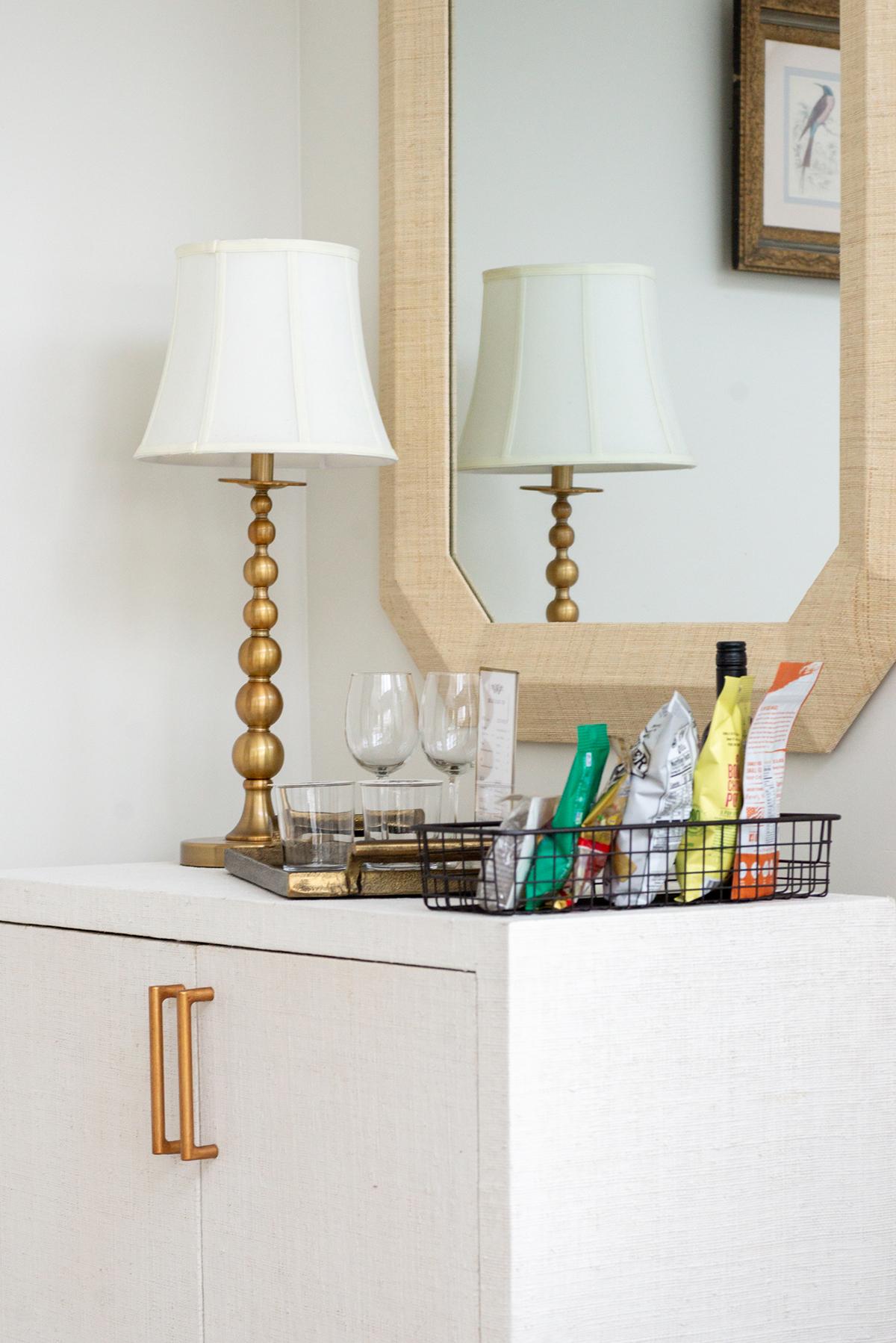 Golden lamp, mirror, snacks, and wine glasses on beige cabinet.