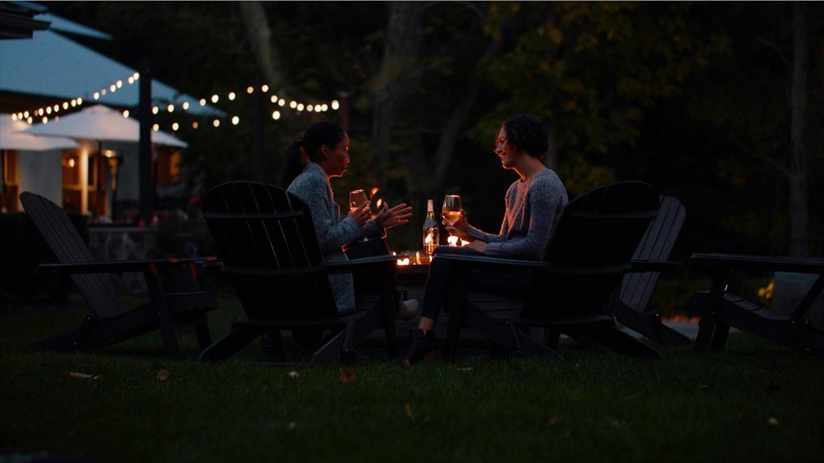 Two people relax by a fire pit at night with string lights in the background.