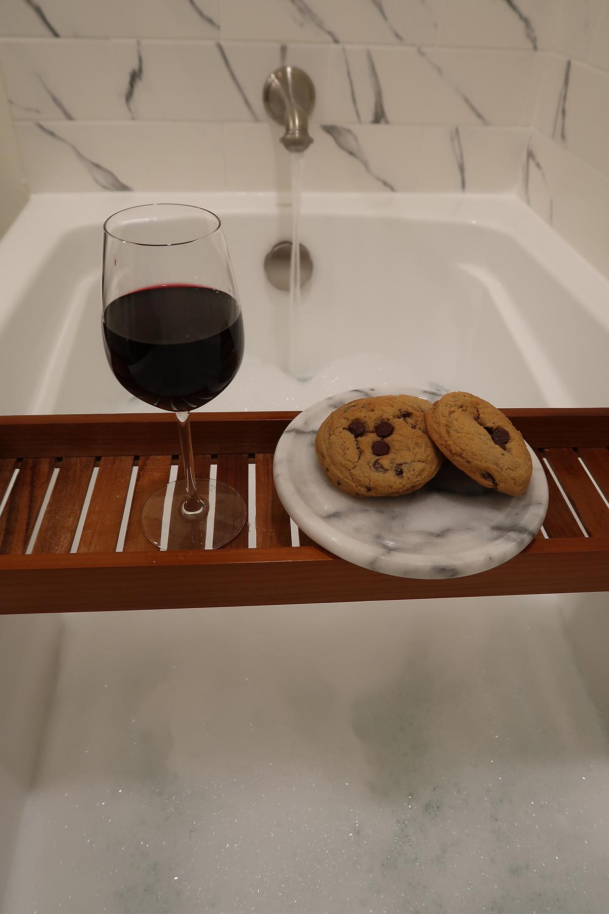 A wine glass and cookies on a tray above a bathtub.