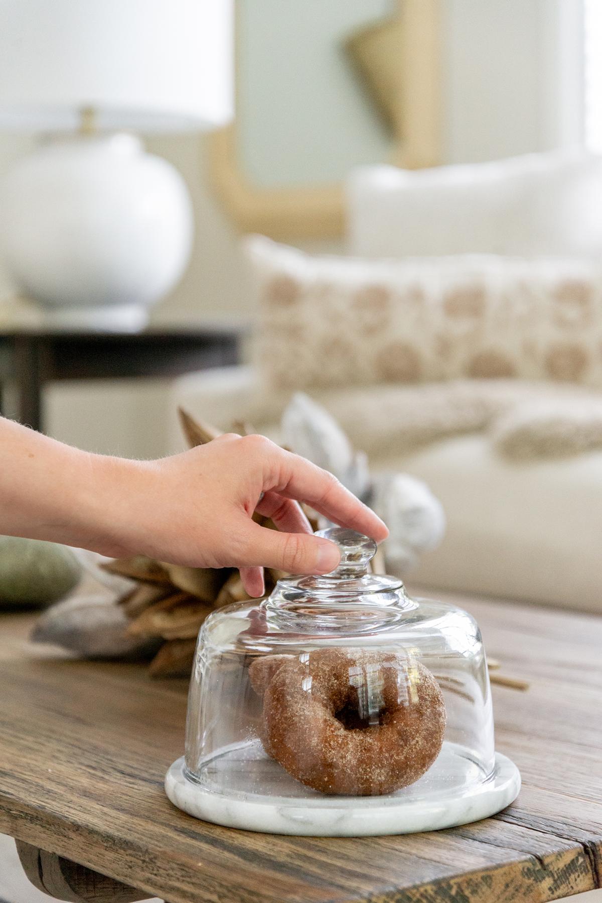 Hand lifting a glass dome covering a donut on a wooden table.