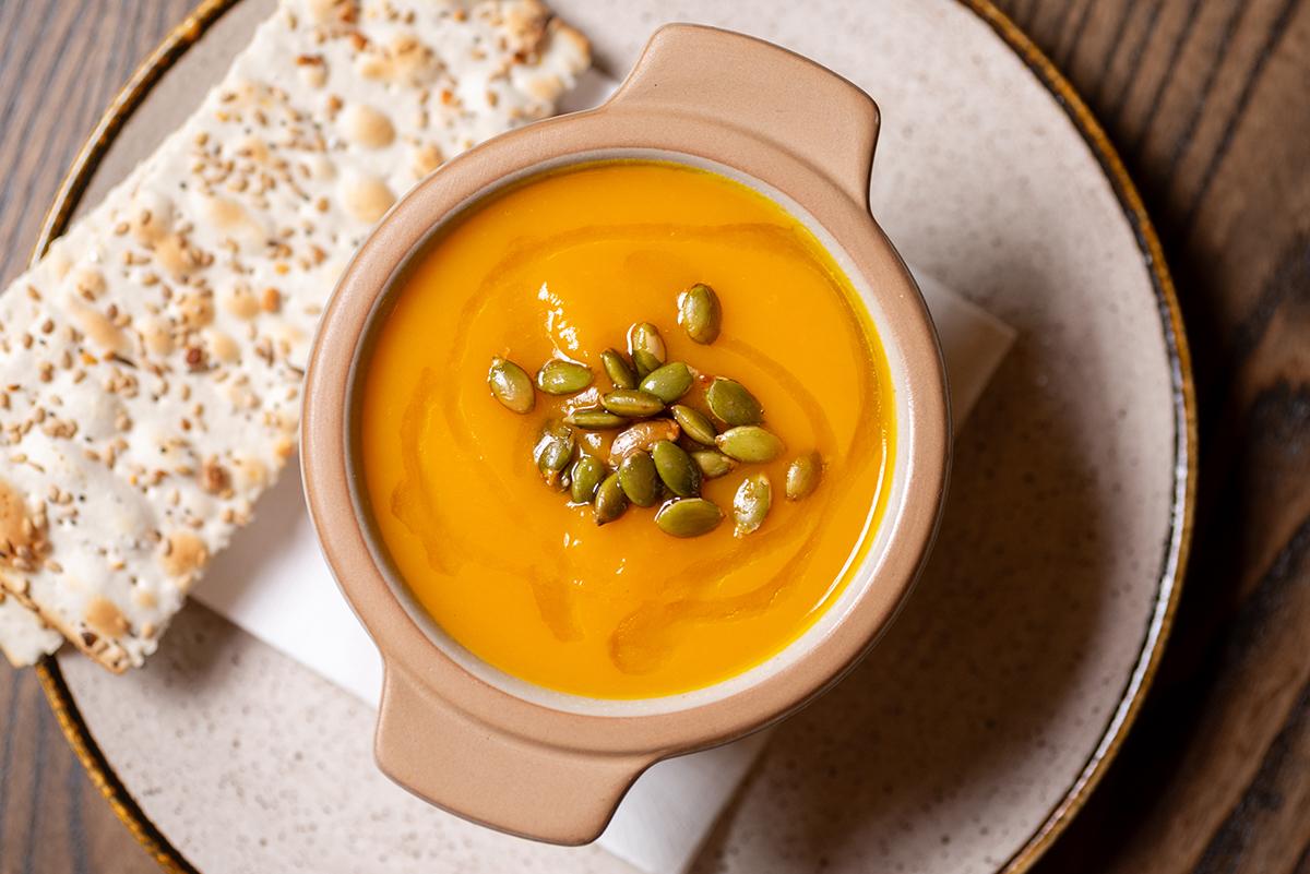 Creamy squash soup garnished with seeds, beside a slice of crispbread on a ceramic plate.