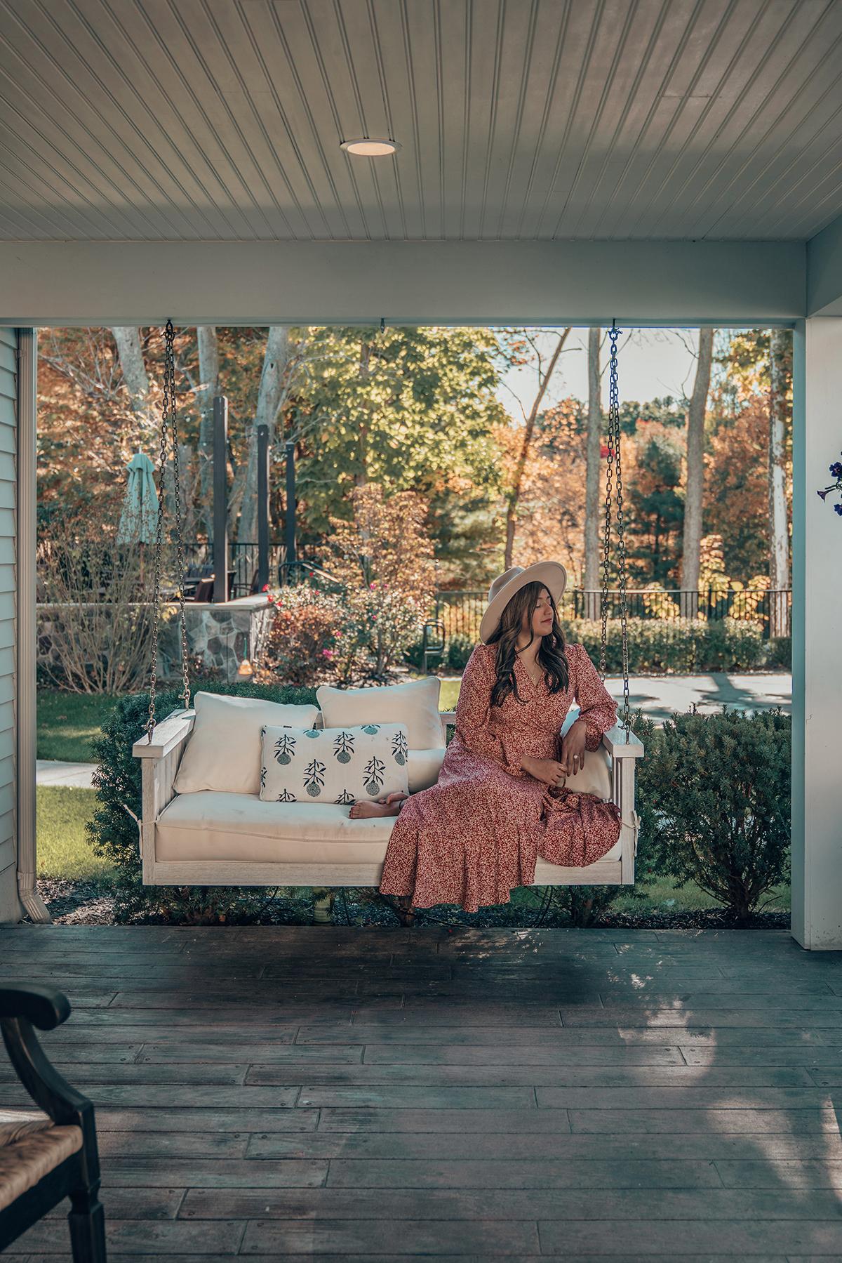Woman in a dress and hat sits on a porch swing in an autumn setting.