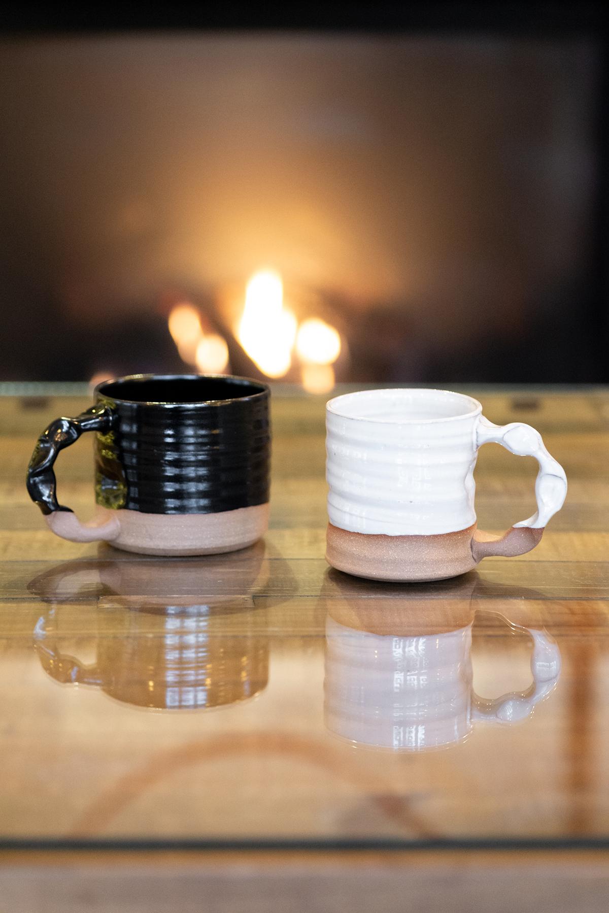 Two ceramic mugs, one black and one white, on a wooden table by a cozy fireplace.