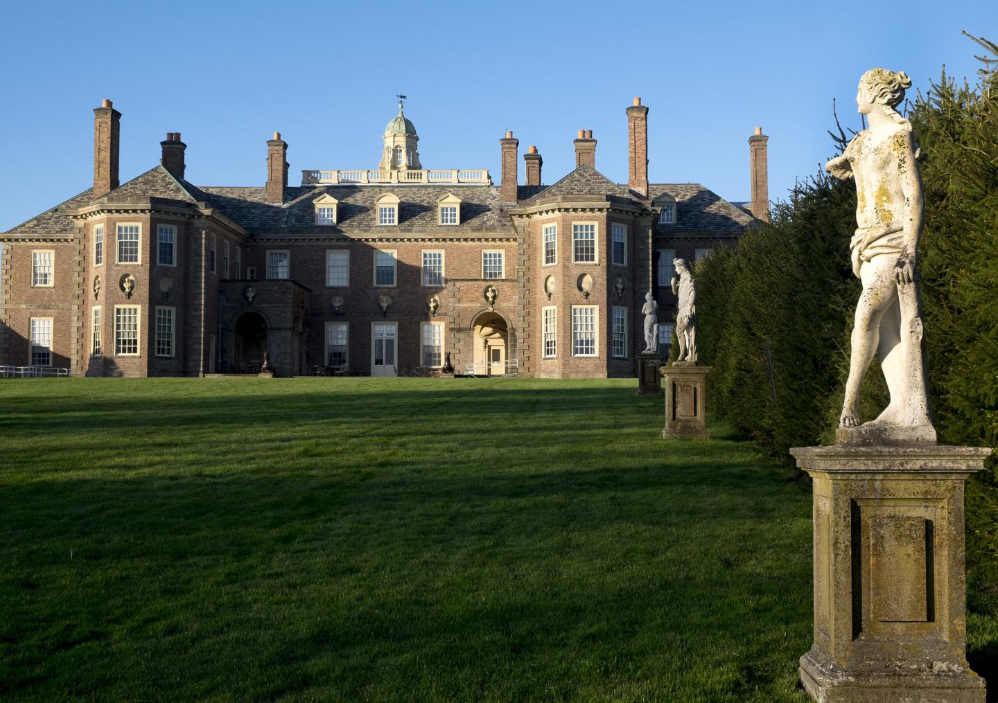 Large brick mansion with statues and green lawn under blue sky.
