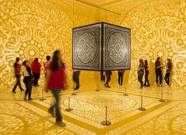 Ornate cube art installation in a yellow-lit room, surrounded by people.