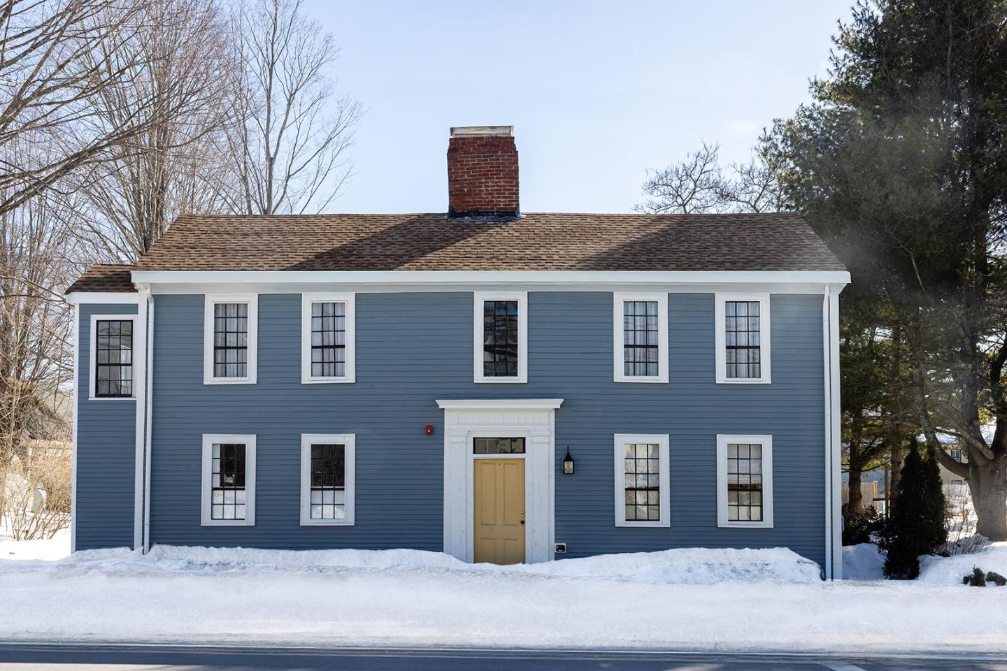 Blue cottage with chimney, snow-covered yard, and bare trees in winter.
