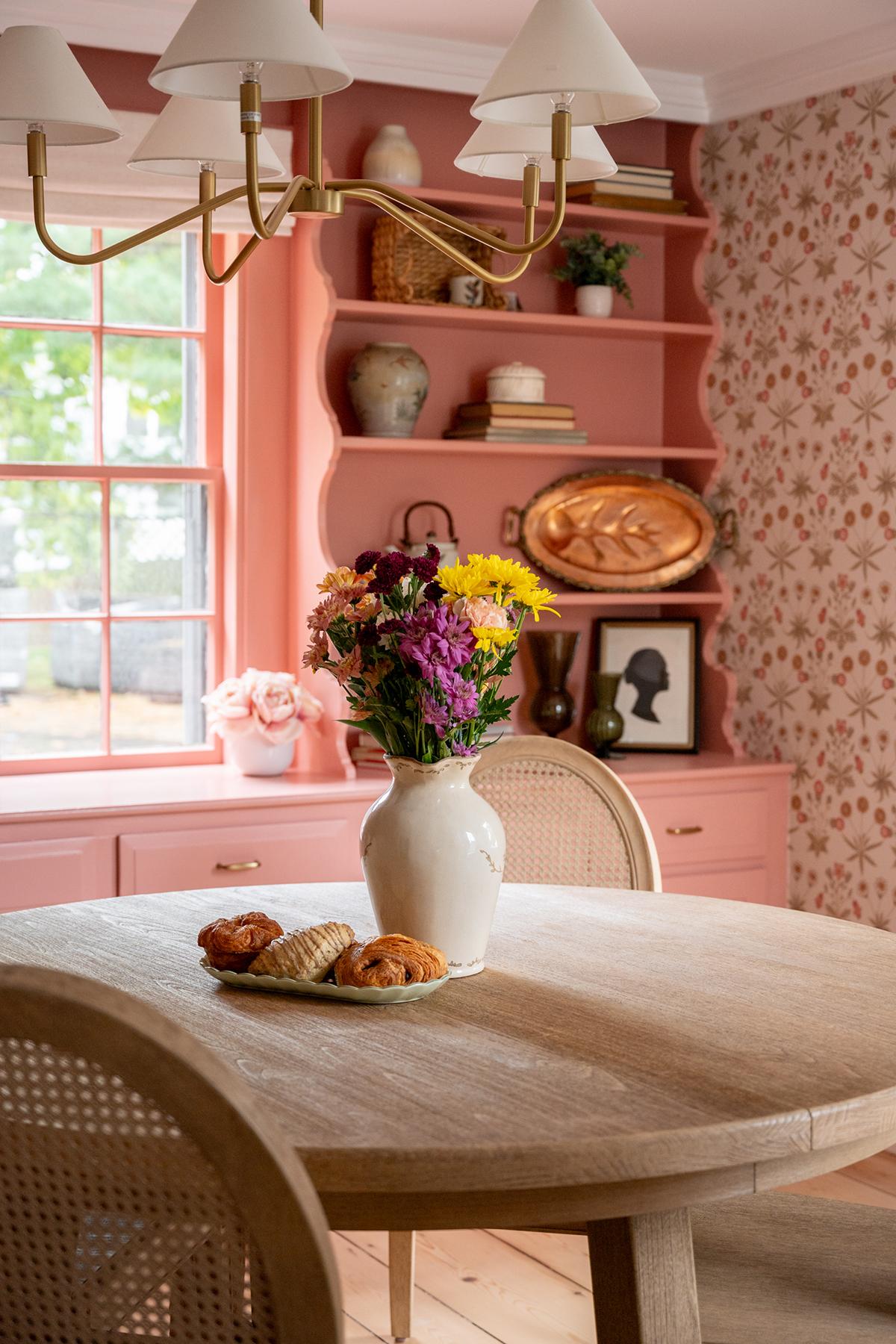 Dining room with pink decor, flowers in vase, and pastries on table.