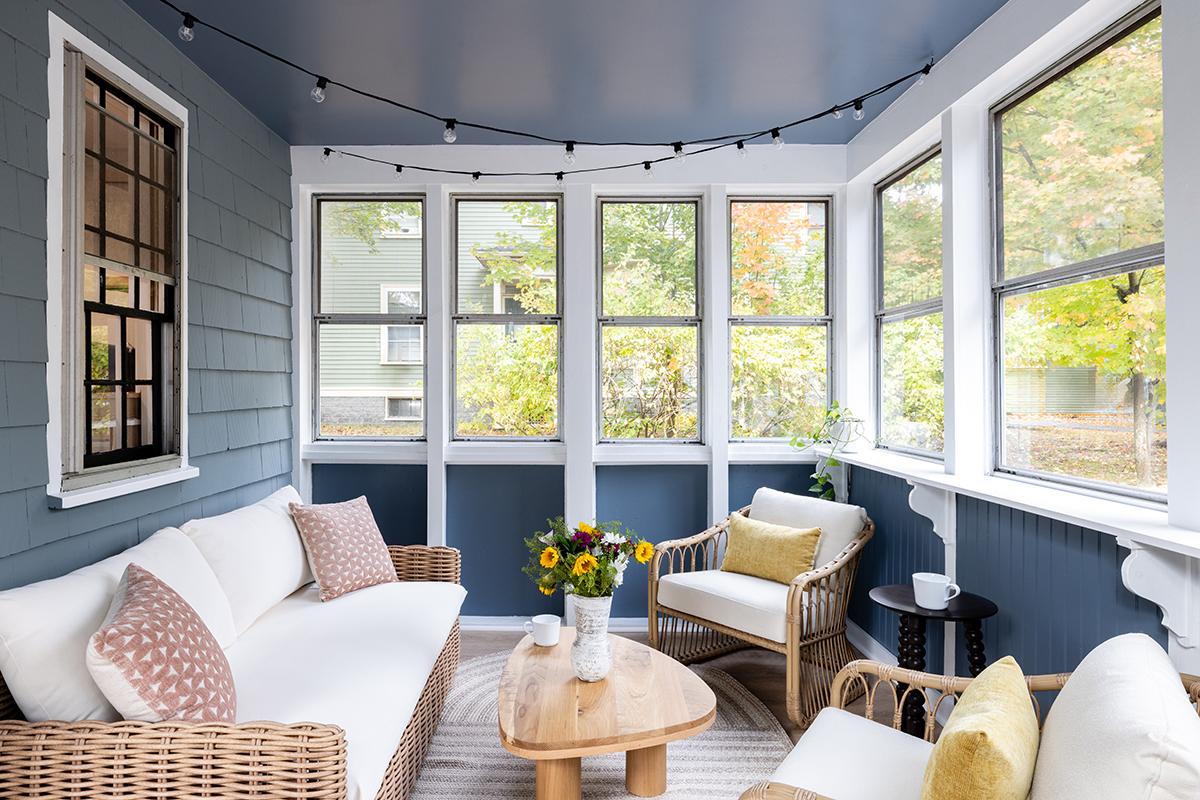 Cozy sunroom with a white sofa, wicker chairs, and a wooden table. Large windows overlook trees.