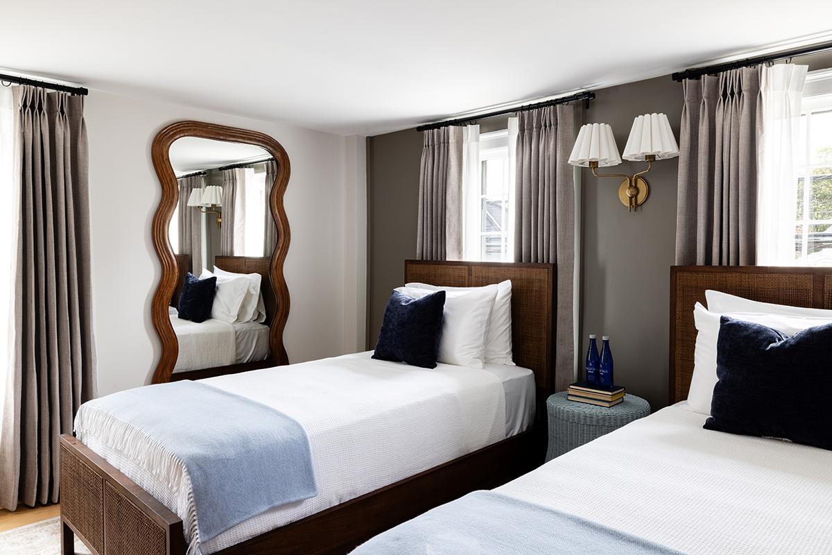 Two neatly made twin beds with blue accents in a bright bedroom with a large mirror.