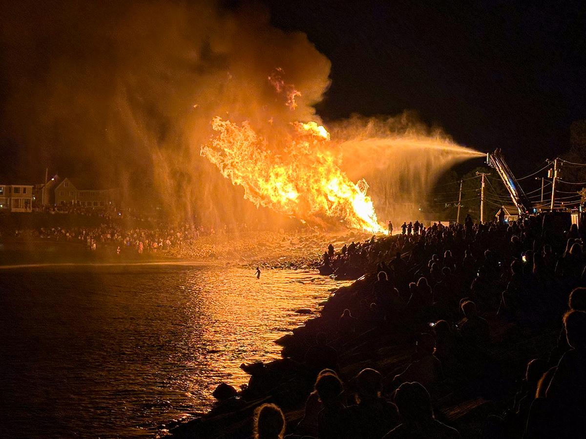 Large bonfire at night with crowd watching by a riverbank.