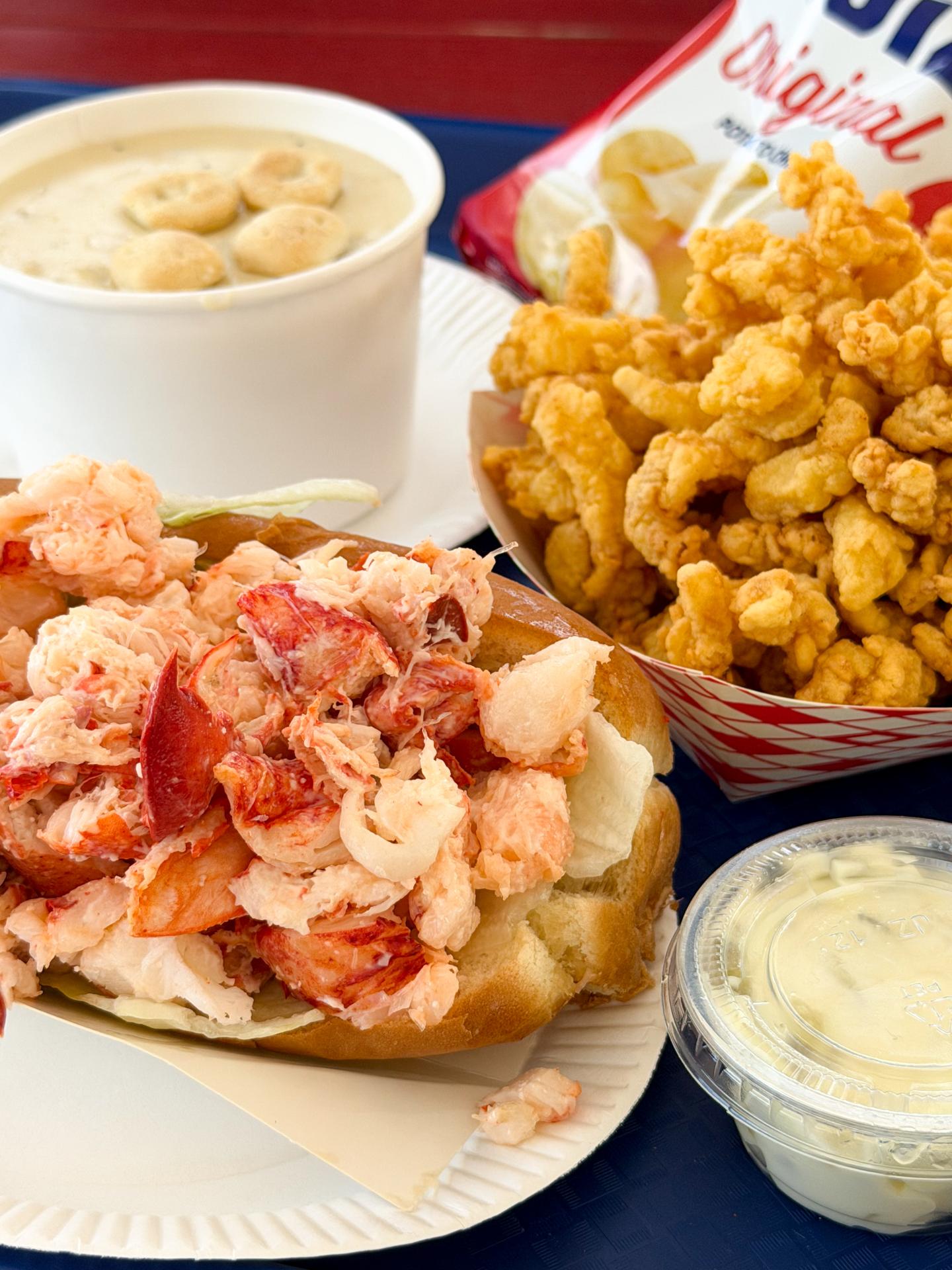 Lobster roll, clam chowder, fried clams, and sauce on a table.