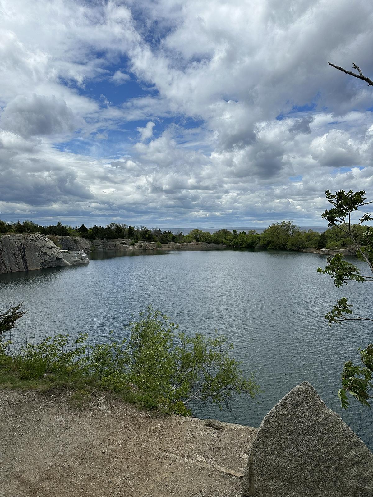 Cloudy sky over a calm lake surrounded by greenery and rocky shore.