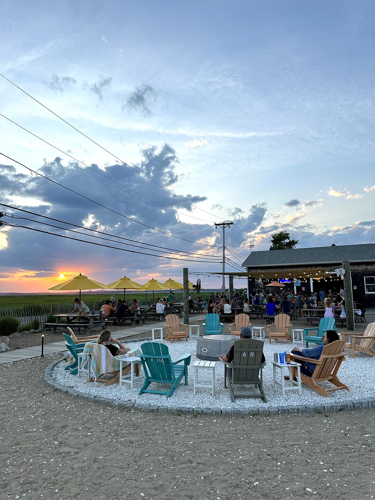 Outdoor seating area at sunset with chairs and people, by a bar under a cloudy sky.