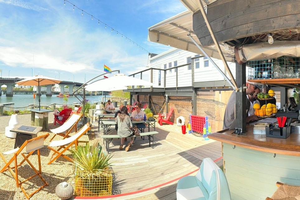 Outdoor cafe by the water with tables, umbrellas, and a bar under a sunny sky.