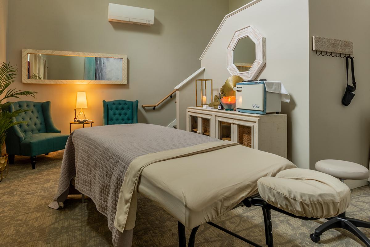 Massage room with a treatment table, warm lighting, and teal chairs.