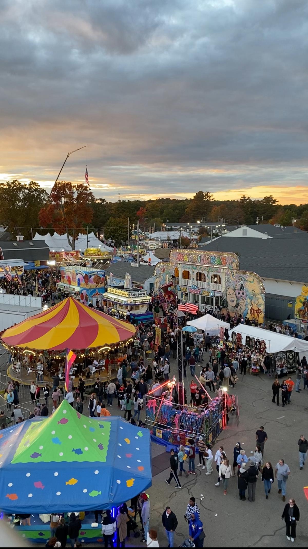 Crowded evening carnival with colorful rides, tents, and a Ferris wheel under cloudy skies.