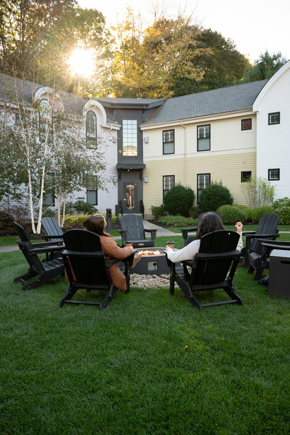Three people relax in Adirondack chairs around a fire pit in a backyard.