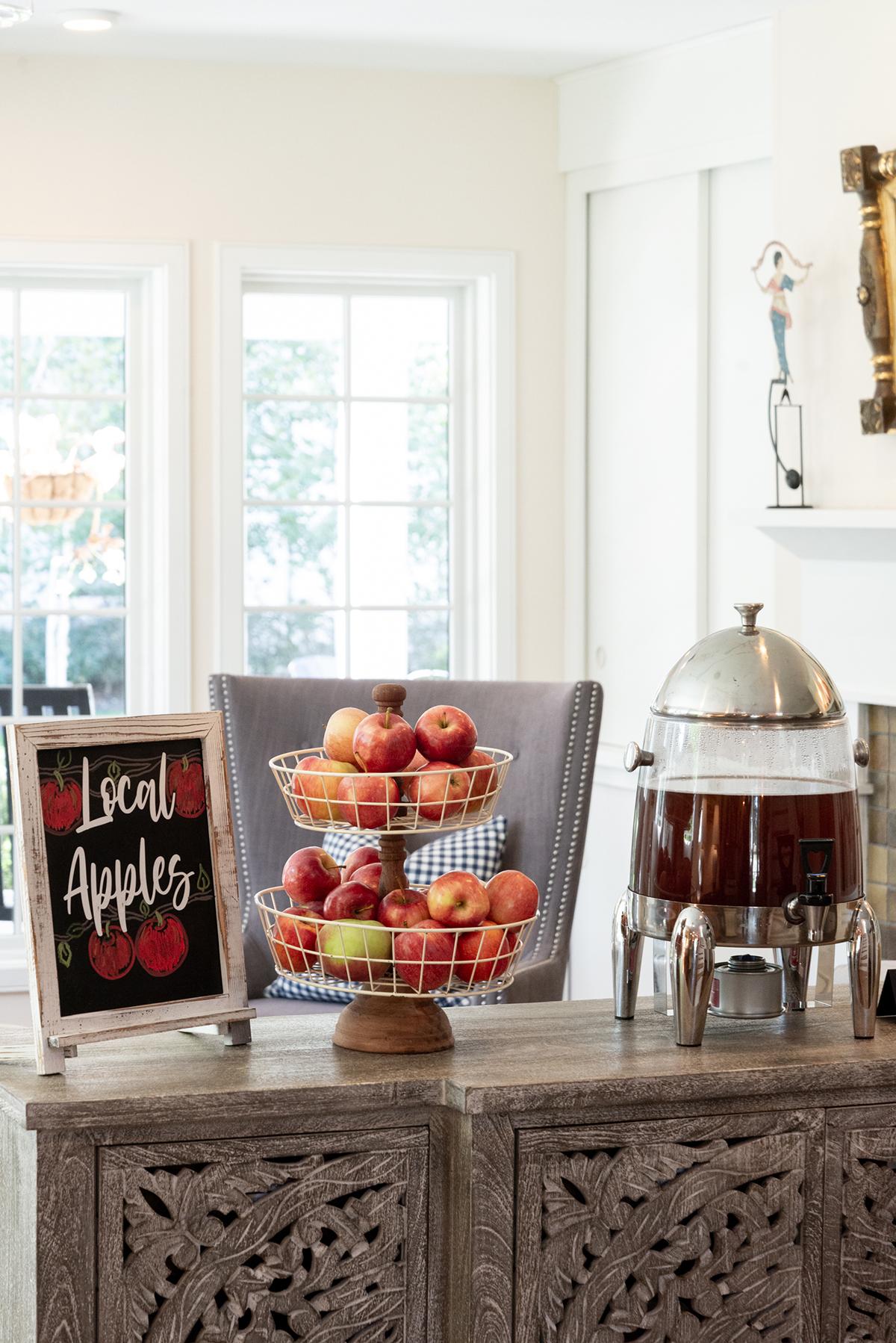 Bright kitchen buffet with tiered fruit stand, coffee dispenser, and “You Are Apple” sign.