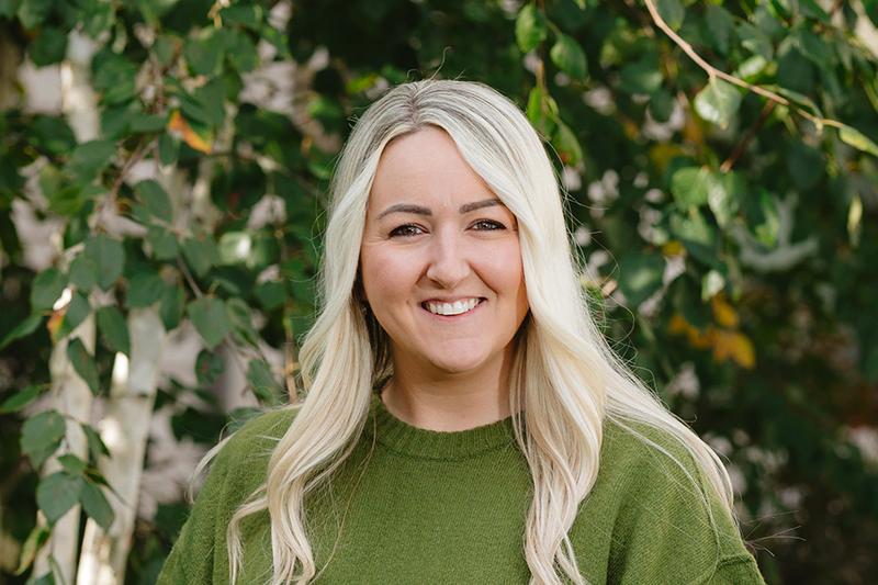 Smiling blonde woman in a green sweater outdoors among leafy trees.