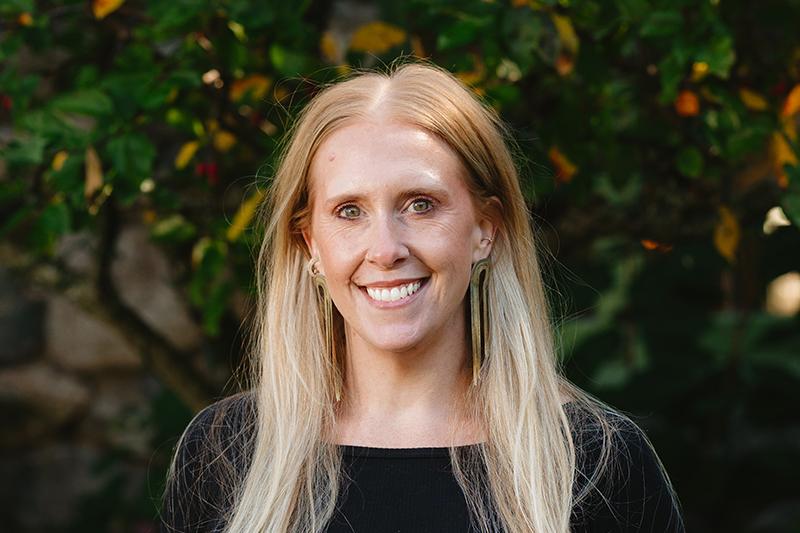 Portrait of a smiling blonde woman outdoors against leafy greenery.