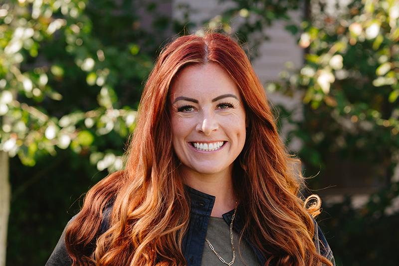 Smiling red-haired woman outdoors with leafy greenery behind her.