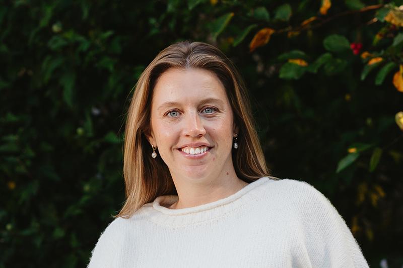 Smiling woman in a white sweater outdoors against leafy greenery
