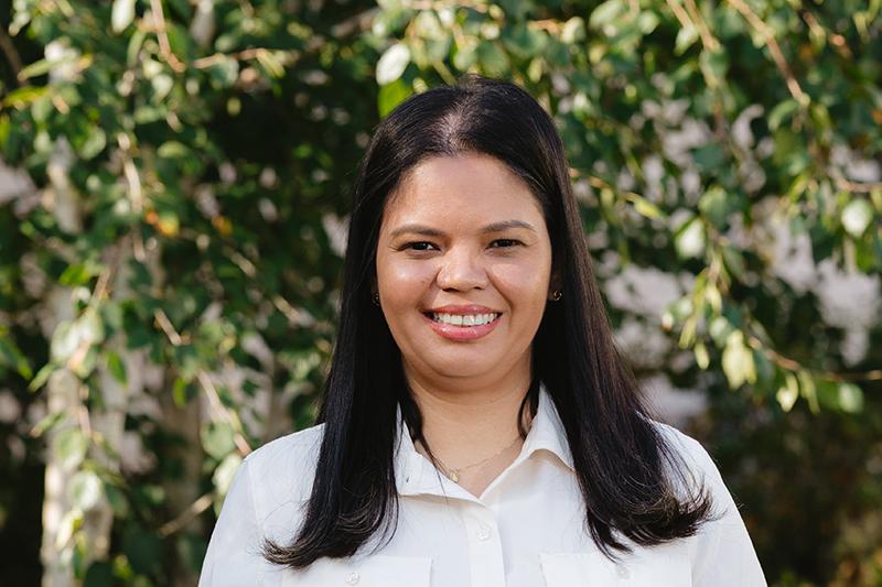 Smiling woman in a white blouse standing outdoors before leafy greenery.